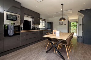 A modern kitchen with dark cabinetry, a built-in microwave, and a wooden dining table with chairs. - Riverside lodge with spacious decking, Ninja BBQ, Wi-Fi, Netflix-ready TV, EV charging and an outdoor copper bath and shower for a wellness experience in nature. Up to three well-behaved dogs are welcome in selected lodges. (Cornwall)