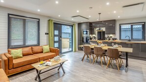 A modern living room with a sofa, coffee table, and dining area with chairs and a table. - Riverside lodge with spacious decking, Ninja BBQ, Wi-Fi, Netflix-ready TV, EV charging and an outdoor copper bath and shower for a wellness experience in nature. Up to three well-behaved dogs are welcome in selected lodges. (Cornwall)
