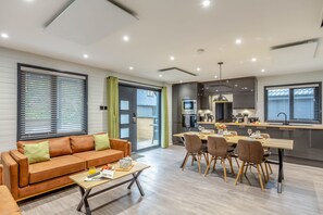 A modern living room with a sofa, coffee table, and dining area with chairs and a table. - Riverside lodge with spacious decking, Ninja BBQ, Wi-Fi, Netflix-ready TV, EV charging and an outdoor copper bath and shower for a wellness experience in nature. Up to three well-behaved dogs are welcome in selected lodges. (Cornwall)