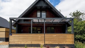 A modern house with a sloped roof, wooden exterior, and a covered porch area. - Riverside lodge with spacious decking, Ninja BBQ, Wi-Fi, Netflix-ready TV, EV charging and an outdoor copper bath and shower for a wellness experience in nature. Up to three well-behaved dogs are welcome in selected lodges. (Cornwall)