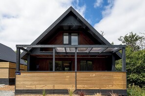 A modern house with a sloped roof, wooden exterior, and a covered porch area. - Riverside lodge with spacious decking, Ninja BBQ, Wi-Fi, Netflix-ready TV, EV charging and an outdoor copper bath and shower for a wellness experience in nature. Up to three well-behaved dogs are welcome in selected lodges. (Cornwall)