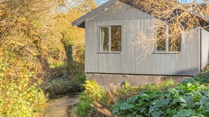 Une petite cabane rustique, avec un toit en métal, entourée d’une végétation luxuriante et bordée d’un ruisseau étroit.