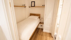 Une petite chambre avec un lit simple, une table de chevet, une commode, un tableau encadré au mur et une étagère en bois avec de petites plantes.