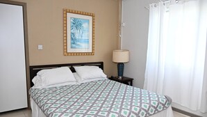 A bedroom with a bed, a nightstand, a lamp, a framed picture, and a window with curtains. - Spacious 2BR Apartment in Cap Estate, Saint Lucia - Walk to Beaches (Gros Islet)