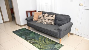 A grey sofa with two decorative pillows, a green patterned rug, and a wooden side table in a room with white walls and a tiled floor. - Spacious 2BR Apartment in Cap Estate, Saint Lucia - Walk to Beaches (Gros Islet)