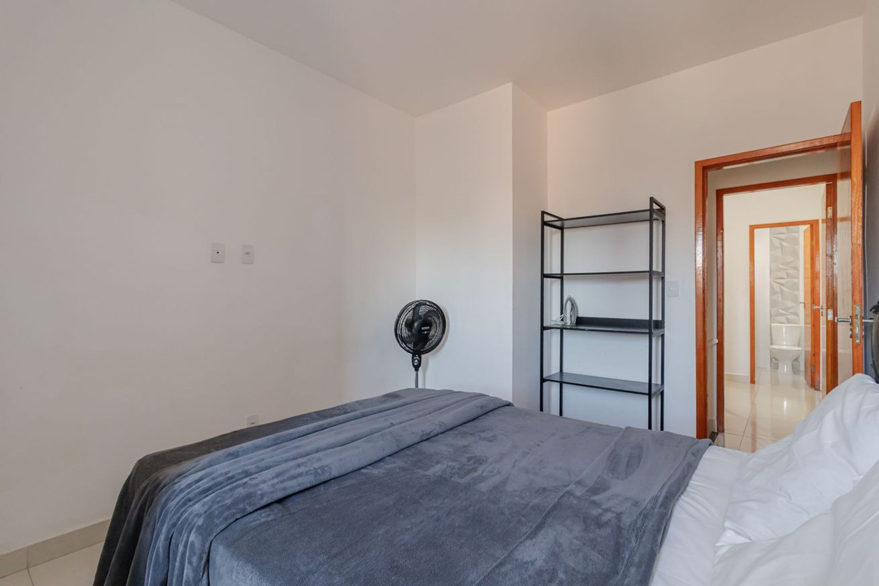 A bedroom with a bed, a standing fan, and a shelving unit.