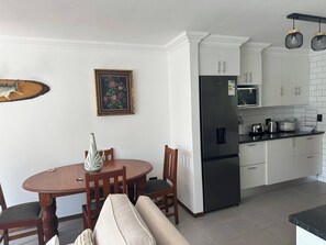 A modern kitchen with white cabinets, a stainless steel refrigerator, a microwave, and a dining table with chairs. - Rosarios Village 2-bed Unit 01, Bryanston (Johannesburg)