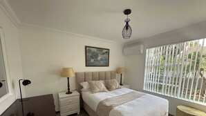 Een slaapkamer met een bed, nachtlampjes, een schilderij, een airconditioner en een raam met jaloezieën.