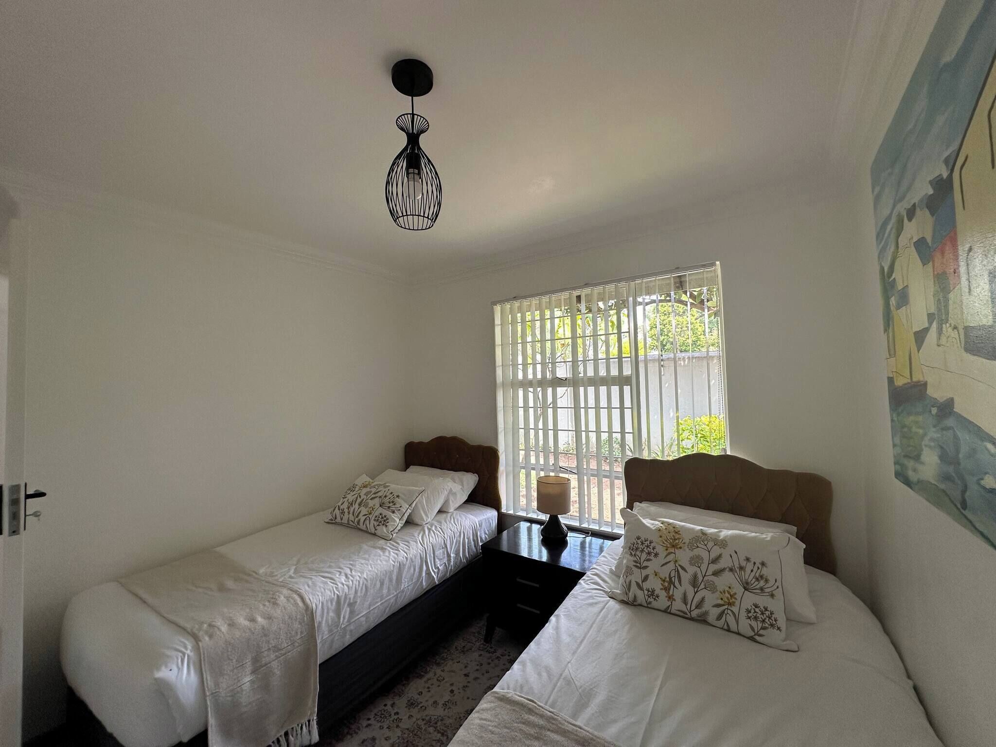 Ein Zimmer mit zwei Betten, einer Pendelleuchte, einem Fenster mit Jalousien und einem Teilbild an der Wand.