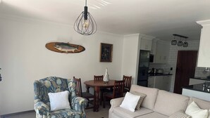 Een woonkamer met een gestreept fauteuil, een beige bank, een eettafel en een keuken op de achtergrond.