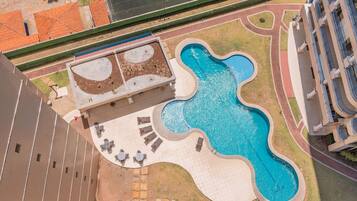 Vista aérea de uma piscina com um design ondulado singular, cercada por espreguiçadeiras e uma pequena área ajardinada.