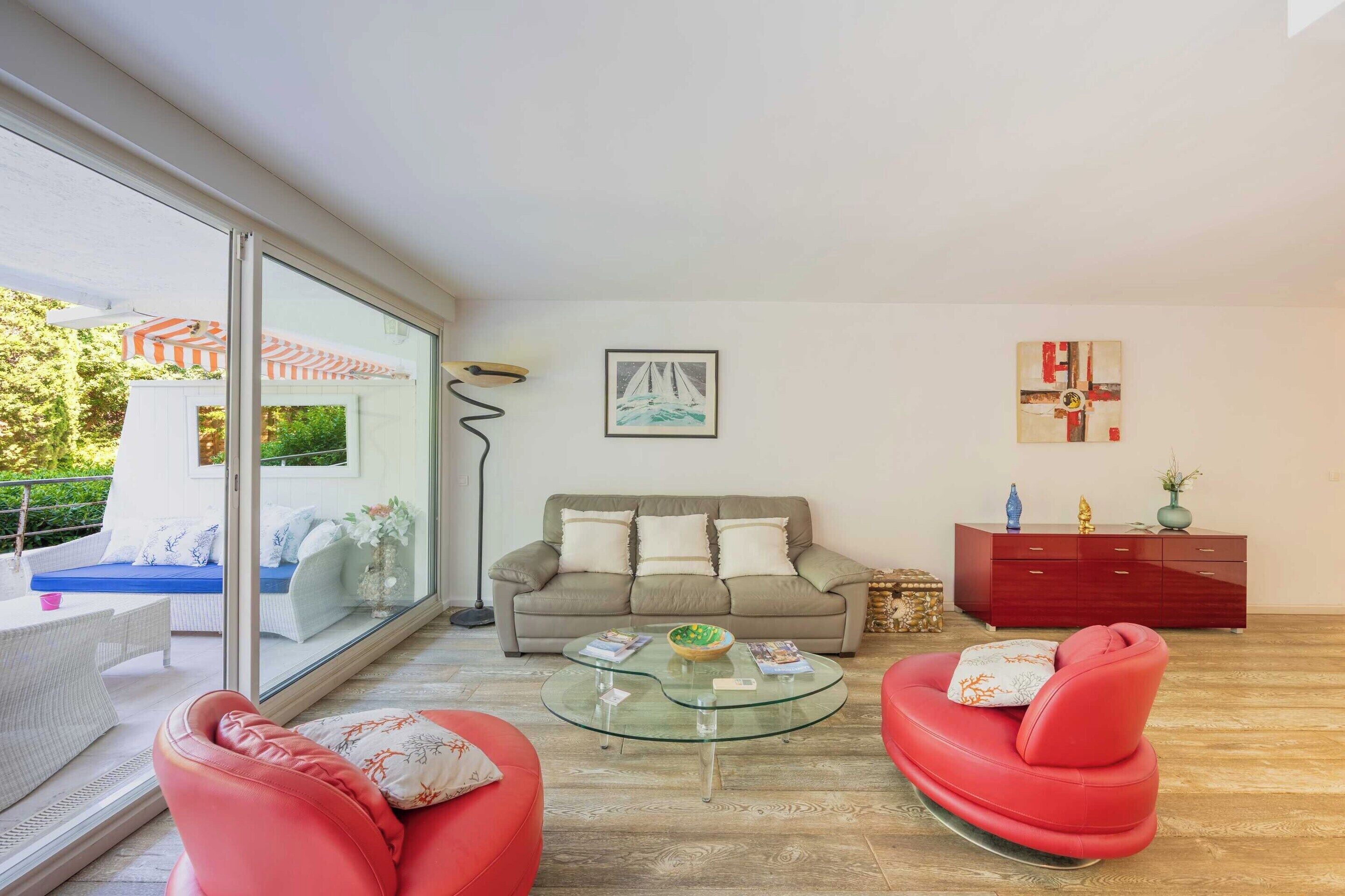 Una sala de estar con una mesa de centro de vidrio, sofás de cuero rojo, un sofá beige, un aparador rojo y un cuadro de un velero en la pared.