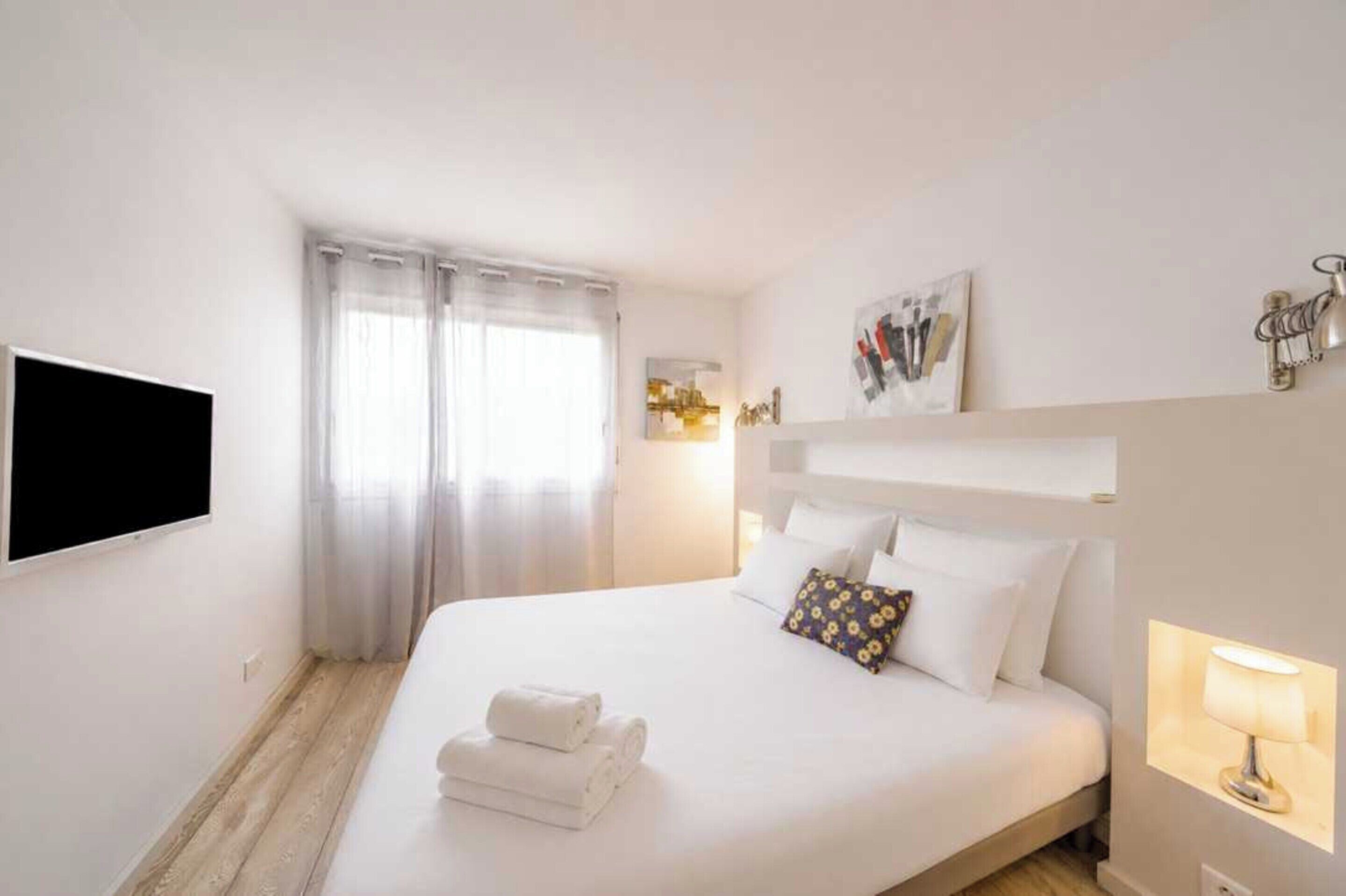 Habitación de hotel con cama, televisor de pantalla plana, ventana con cortinas, lámpara de pared y un cuadro.