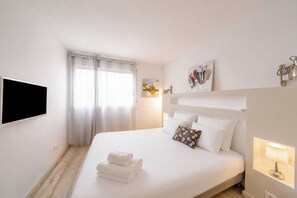 A hotel room with a bed, a flat-screen TV, a window with curtains, a wall-mounted lamp, and a painting. - Charming Apartment With Pool 2br/4p - Saint-tropez (Saint-Tropez)