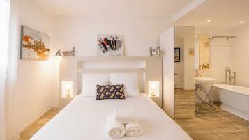 Habitación de hotel con una cama grande, una mesita de noche, un espejo, un baño con bañera y lavamanos.