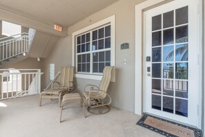 A covered outdoor area with two striped chairs, a white railing, and a door with a window. - White Cap Condo B201 (Fort Myers Beach)