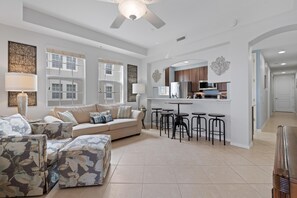 A spacious living room with a sofa, ottoman, and chairs, a kitchen area with bar stools, and large windows. - White Cap Condo B201 (Fort Myers Beach)