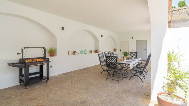 Eine Terrassenanlage im Freien mit Grill, Esstisch, Stühlen, Kübelpflanzen und einer weißen Mauer mit Bögen.