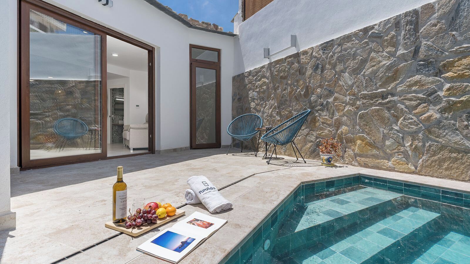 A pool area with a stone wall, a glass door leading inside, and a bottle of wine on the poolside.