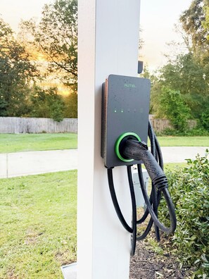An outdoor electric vehicle charging station with a cable plugged in, mounted on a white post.