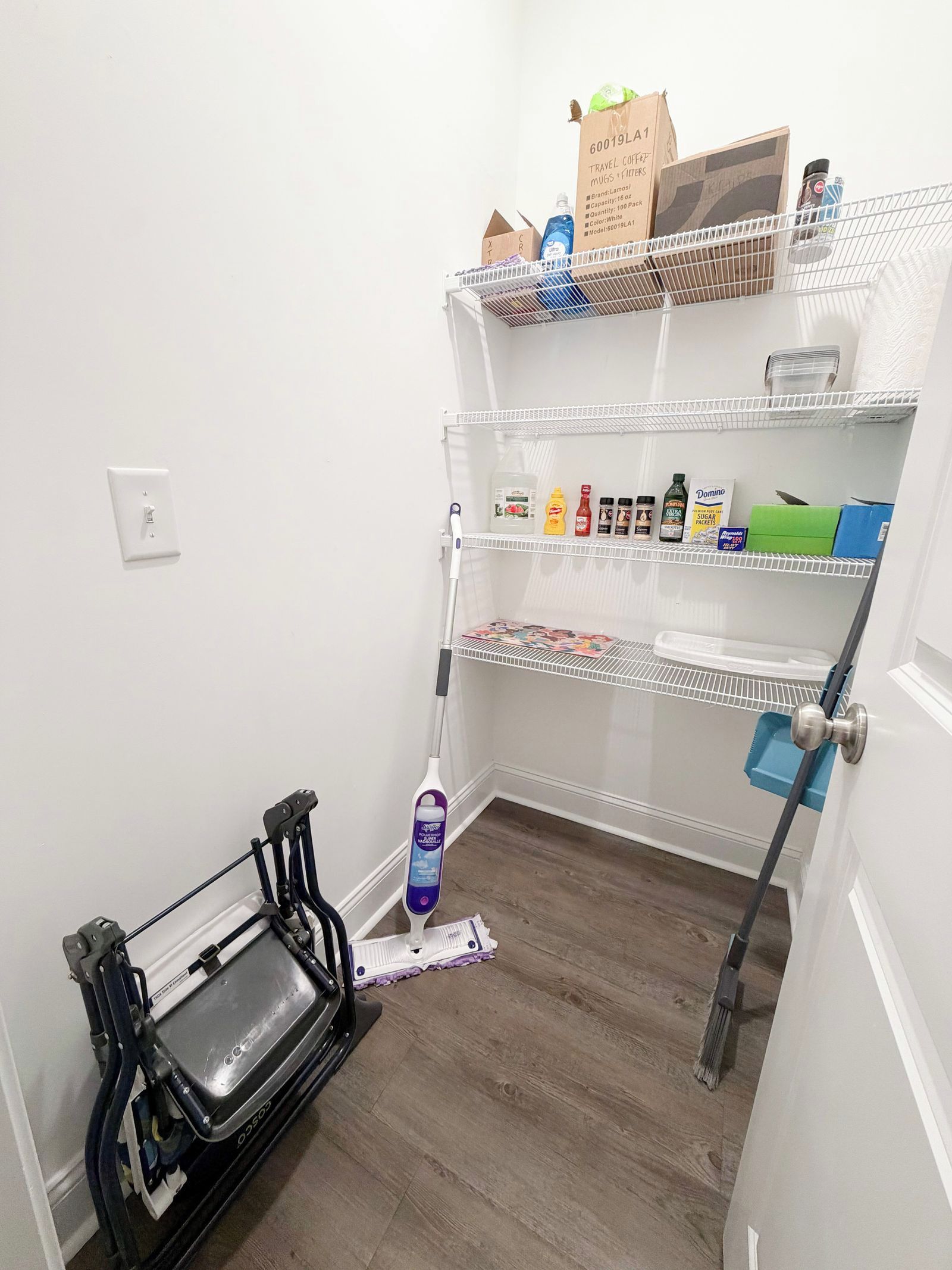 A compact storage area with shelves holding travel mugs, cleaning supplies, and a vacuum cleaner.