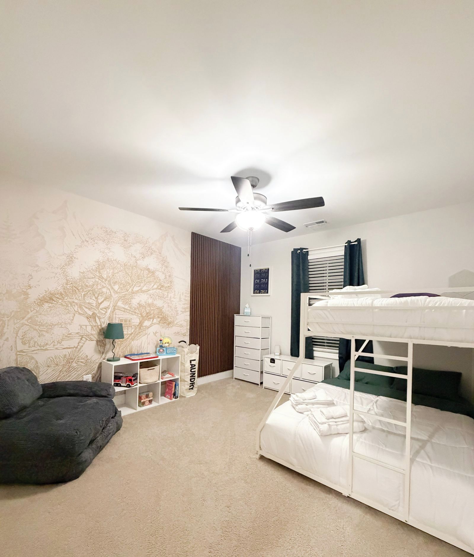 Un dormitorio con literas, ventilador de techo, cómoda, lámpara y pared con un mural de un árbol.