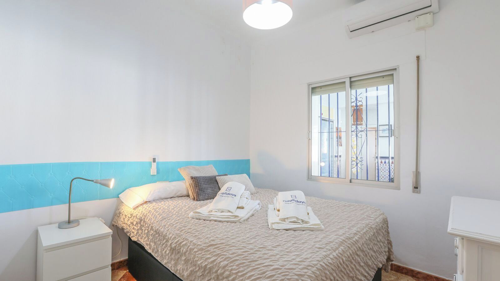 Un lit bien fait, avec un mur d’accent bleu, une lampe de chevet et une fenêtre avec des barreaux.