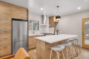 A modern kitchen with a stainless steel refrigerator, wooden cabinets, a white island with bar stools, and a pendant light. - The Madison | Walk to Adams Ave & Cafés (San Diego)