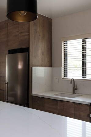 Eine moderne Küche mit einem Edelstahl-Kühlschrank, einer weißen Arbeitsplatte, einem Fenster mit Jalousien und einer Pendelleuchte.