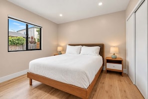 A bedroom with a large bed, two bedside tables with lamps, a window with a view of greenery, and a door to the right. - The Madison | Walk to Adams Ave & Cafés (San Diego)