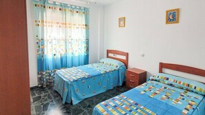 Une chambre avec deux lits, une table de chevet, une tête de lit en bois, une fenêtre avec des rideaux et une petite décoration murale.