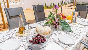 Una mesa de comedor al aire libre dispuesta con platos, cubiertos, una botella de vino, un cuenco de cerezas, pan y un jarrón con flores.