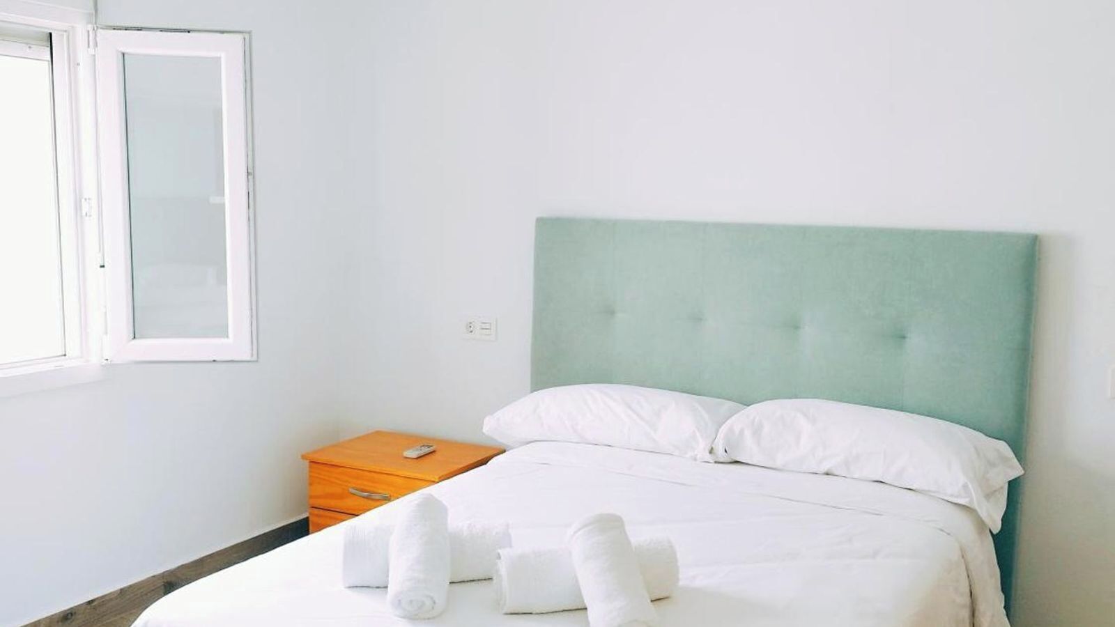 Un lit bien fait, avec du linge de lit blanc, une table de chevet en bois sur laquelle se trouve une télécommande, et une fenêtre à cadre blanc.