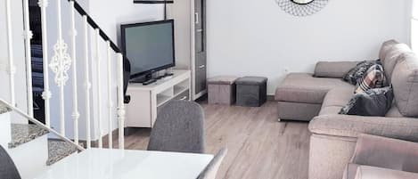 A modern living room with a flat-screen TV, a grey sofa, and a dining area with a white table.