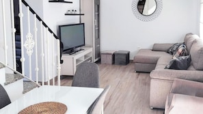 A modern living room with a flat-screen TV, a grey sofa, and a dining area with a white table. - Casa del Tío Paco Montecorto by Ruralidays (Málaga)