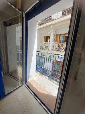 A view through a glass door into a balcony area with a metal railing, a tiled floor, and a window with wooden shutters. - Apartment 'Nou 8' with Balcony, Wi-Fi and Air Conditioning (Sitges)