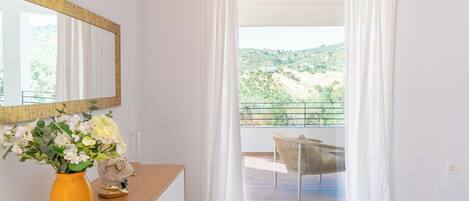 A room with a wooden desk, a vase of flowers, a mirror, and a view of a balcony with a chair.