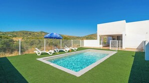 Ein Swimmingpool mit Kunstgraseinsatz, weißen Liegestühlen, einem blauen Sonnenschirm und einem weißen Gebäude mit einer Tür.