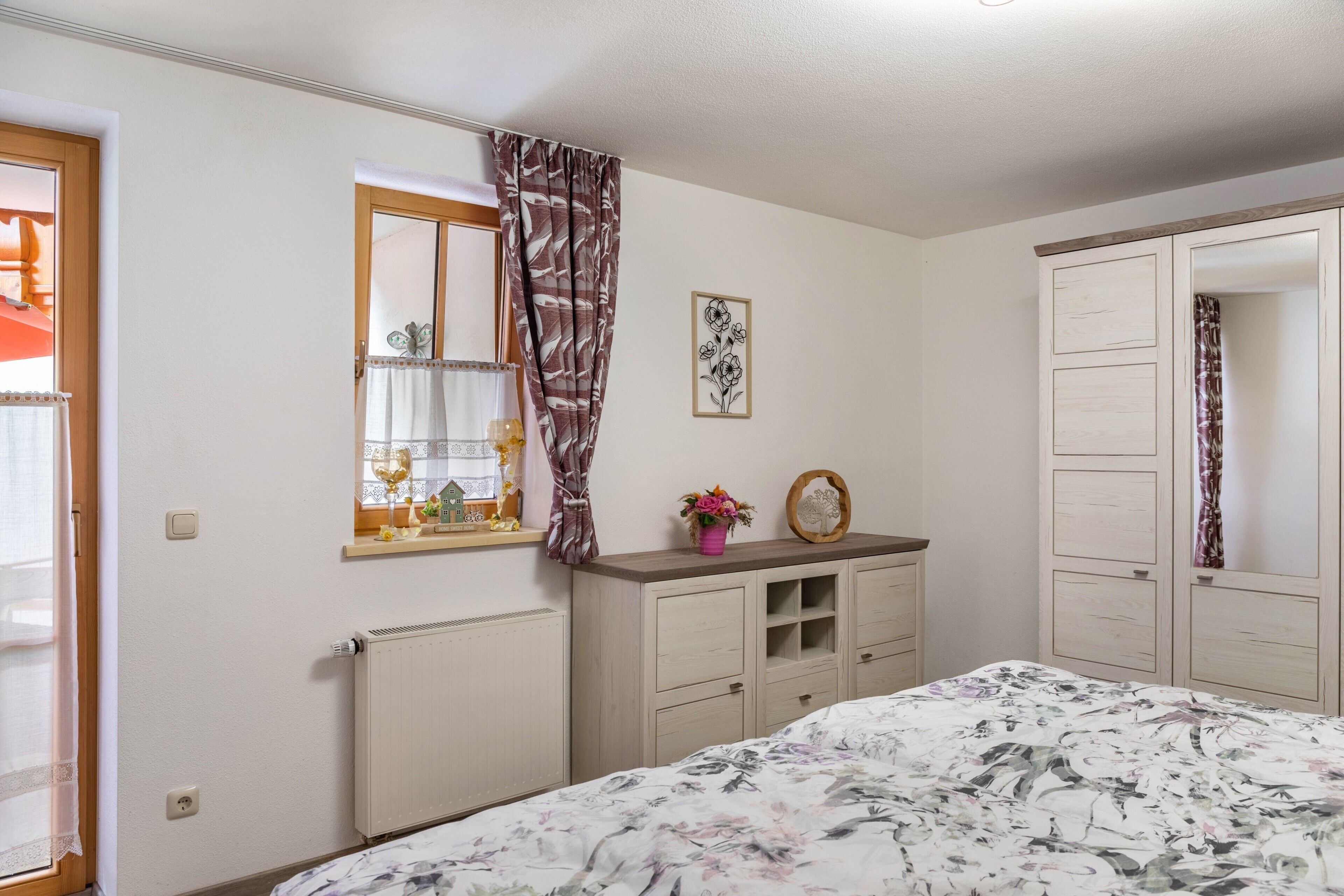 Ein Schlafzimmer mit Bett, Kommode, Spiegel, Fenster mit Vorhängen und Heizkörper.
