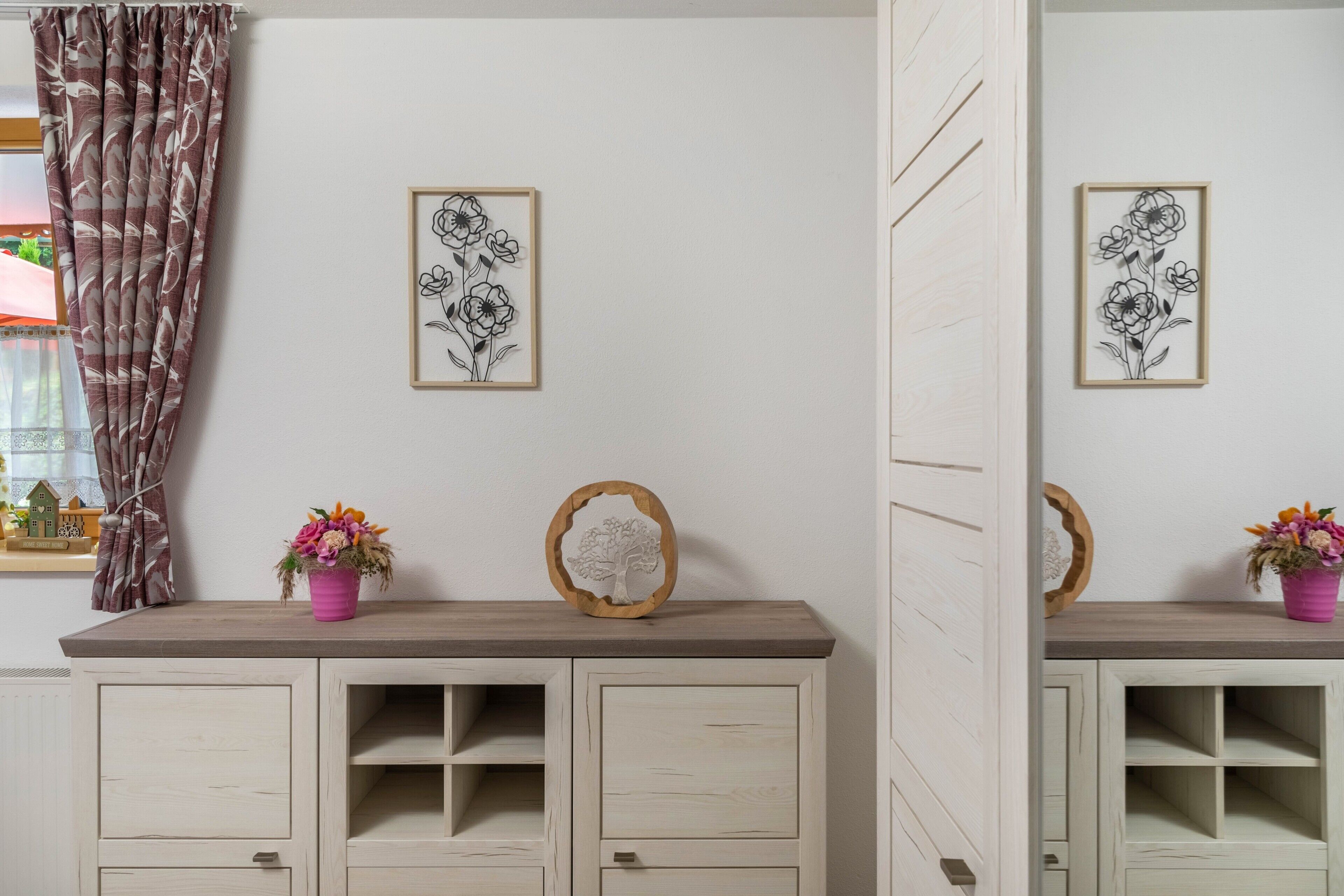 Une pièce comprenant une commode en bois, deux œuvres d’art florales encadrées, un vase rose avec des fleurs et une décoration en bois en forme d’arbre.