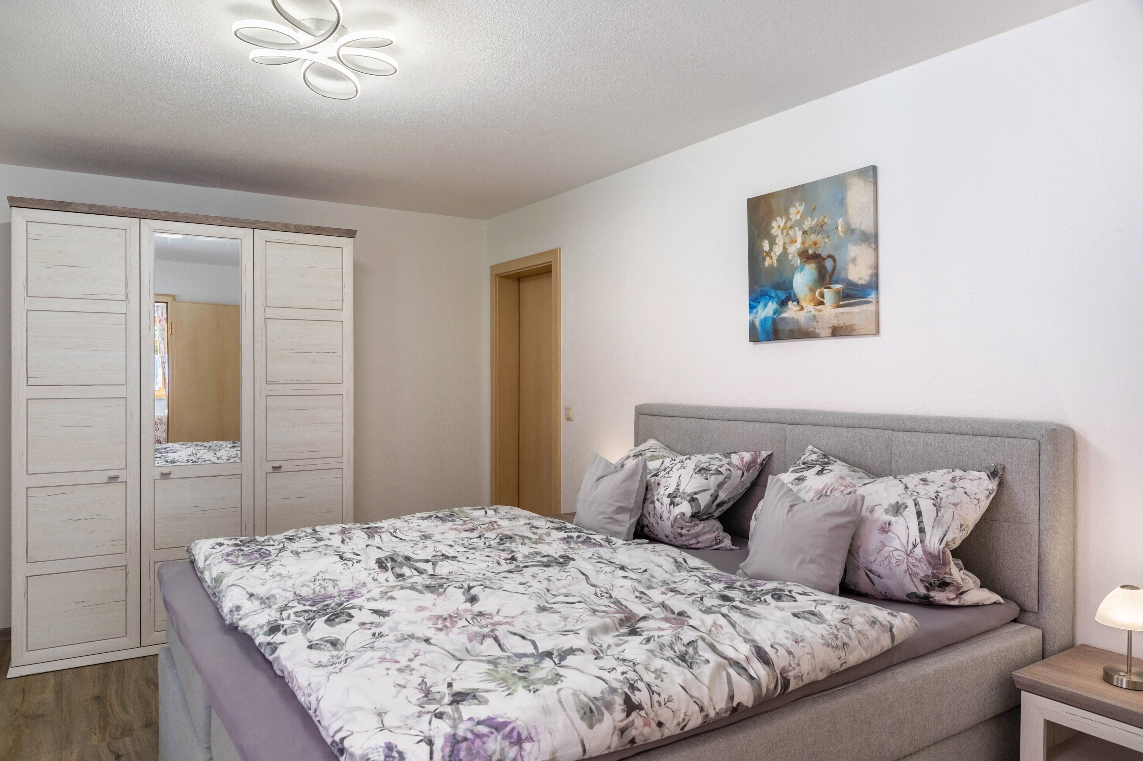 A bedroom with a bed, a nightstand, a lamp, a painting, and a wardrobe.