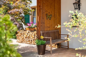 Een houten bank voor een gebouw, met een vlinderdecoratie aan de muur, een bloempot en een stapel houtblokken.