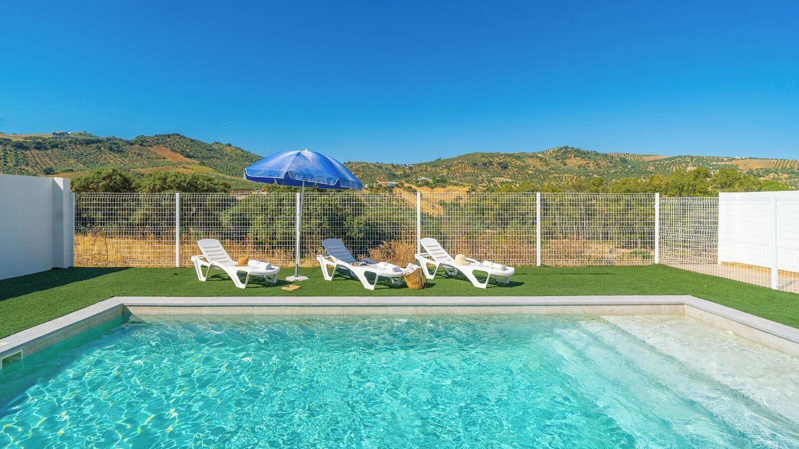 A swimming pool with clear water, surrounded by white lounge chairs and a blue umbrella.