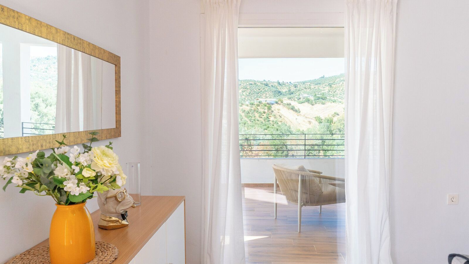 A room with a wooden desk, a vase of flowers, a mirror, and a view of a balcony with a chair.