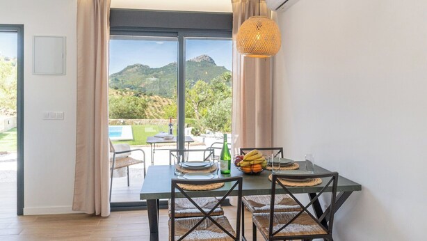 A dining area with a table set for two, a wicker chair, a woven table runner, and a view of a mountainous landscape through a glass door. - Alma de Olivo El Gastor by Ruralidays (Cádiz)