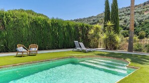 Une piscine dont le bord est recouvert de carrelage, entourée d’une pelouse où se trouvent deux chaises longues et une haie.