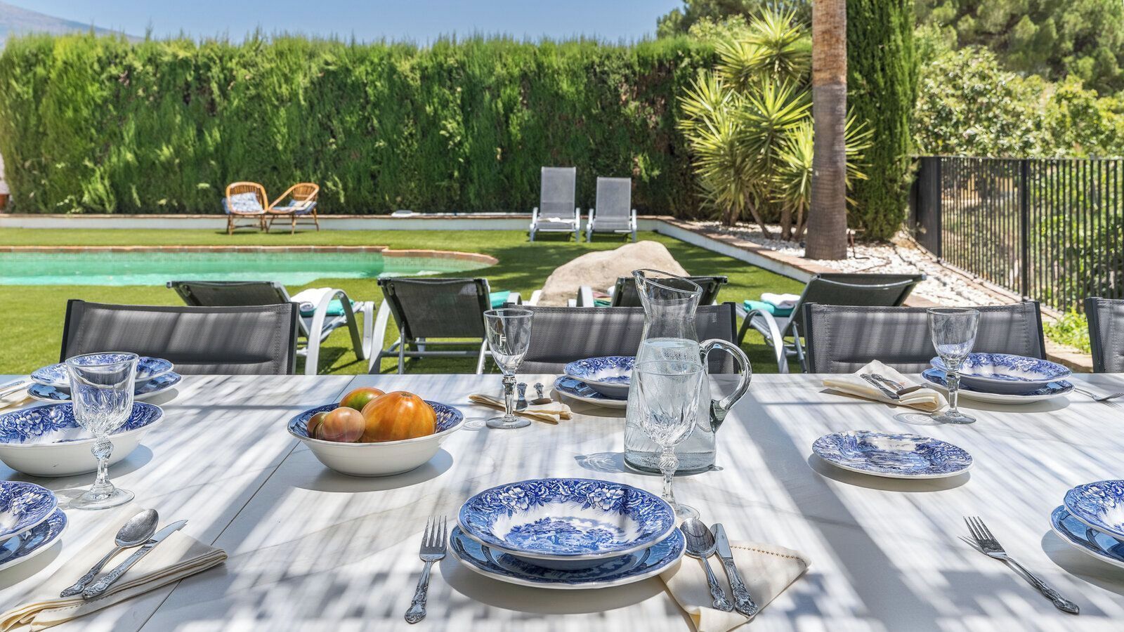 Un espace extérieur aménagé pour les repas, avec une table dressée, entourée de chaises, et une piscine ainsi qu’une végétation luxuriante en arrière-plan.