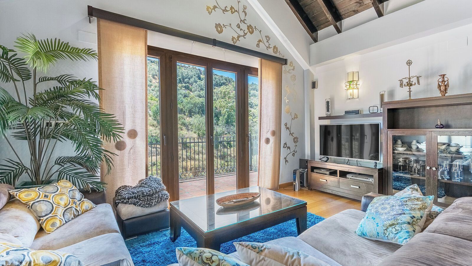 A modern living room with a glass coffee table, a sofa, and a television. There is a balcony with a view of greenery.