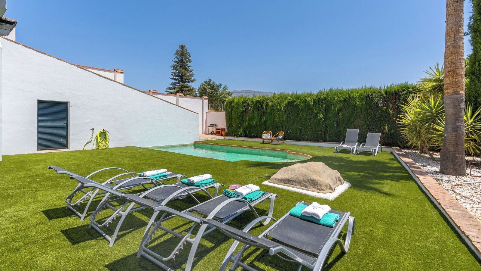 A backyard with a swimming pool, sun loungers, a rock feature, and a white building.