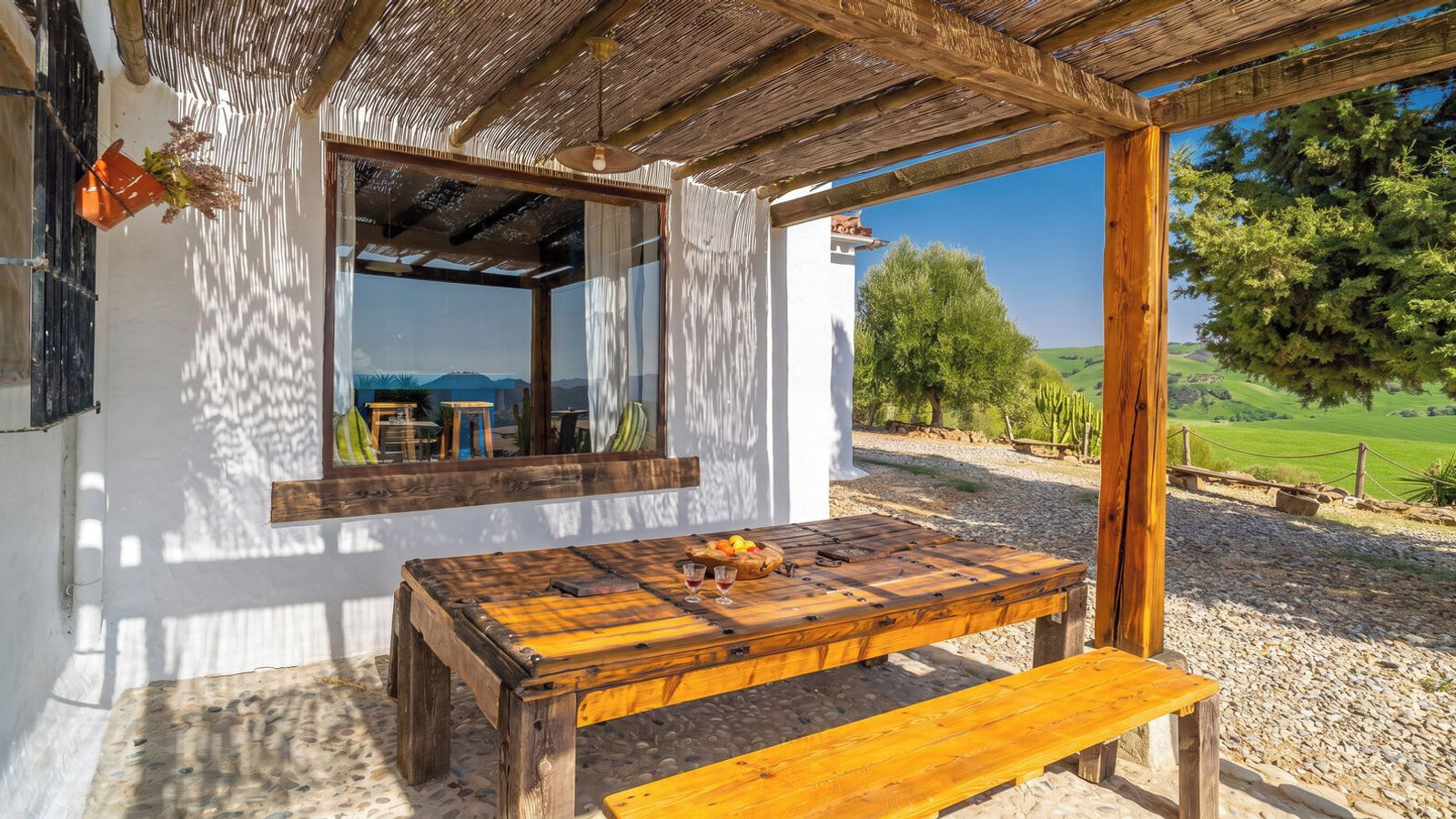 A rustic wooden table with a bench, a hanging orange pot, and a view of a green landscape through an open window.